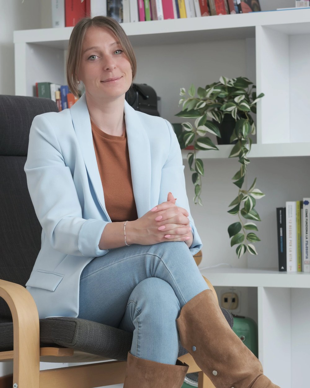 Susanna Manzato in blazer azzurro e stivali marroni su poltrona di legno, davanti a una libreria bianca con pianta