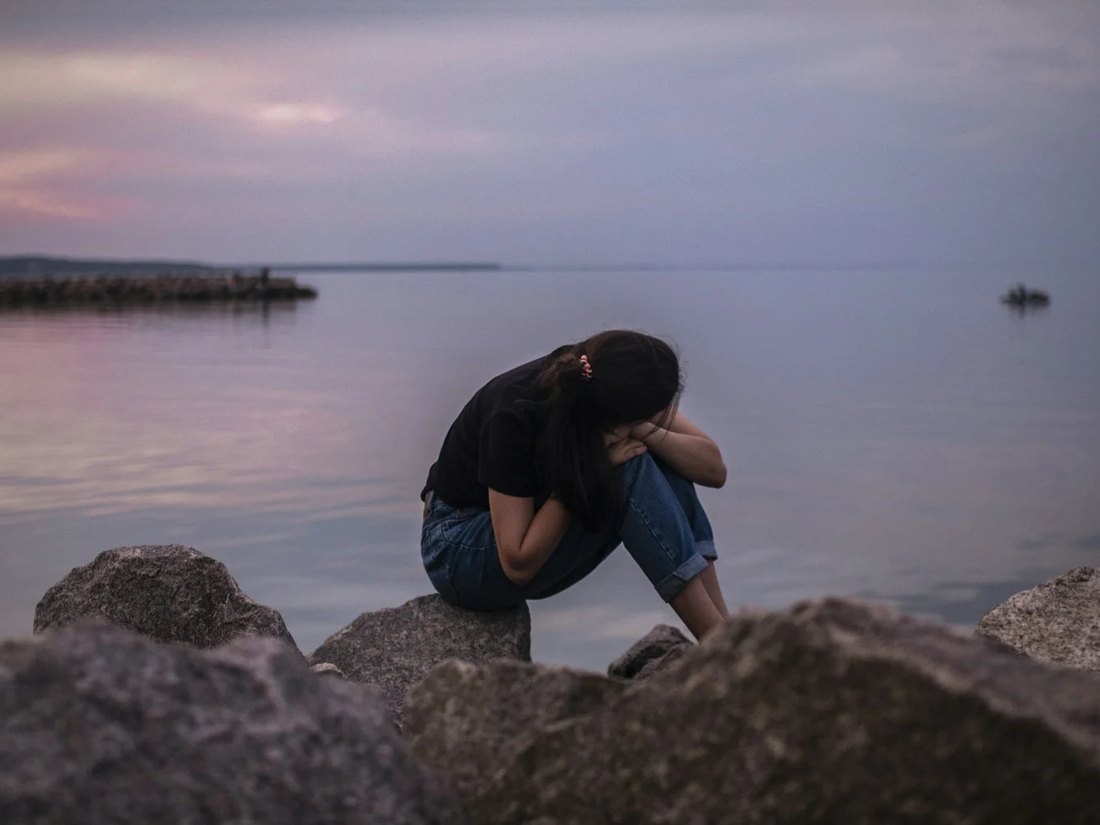 Ragazza con maglietta nera e jeans seduta sugli scogli, testa sulle ginocchia, davanti al lago al tramonto