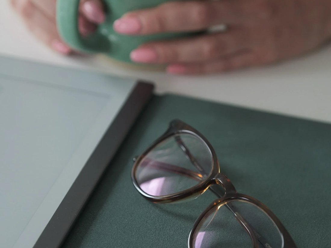 Occhiali con montatura tartarugata su una cartellina verde, sullo sfondo mani con smalto rosa che stringono una tazza verde